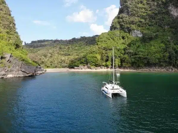 Luftaufnahme eines Katamarans, der in einer ruhigen Bucht vor Anker liegt, umgeben von üppig grünen Klippen und einem abgeschiedenen Sandstrand.