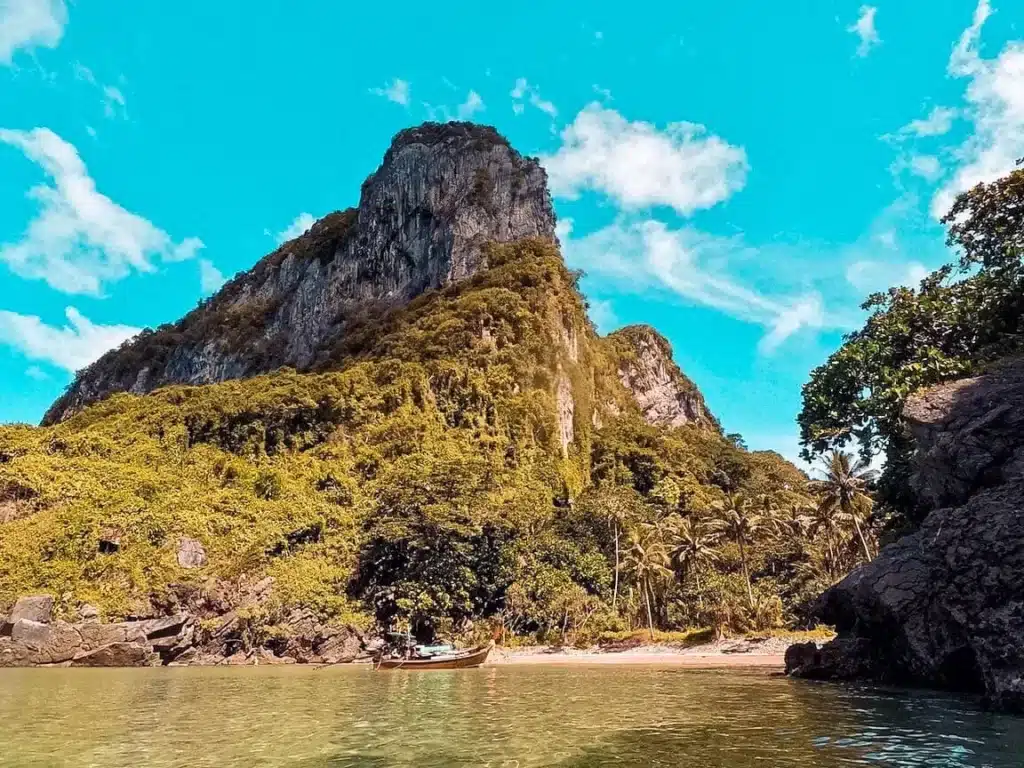 Ein lebendiges Bild, das eine hoch aufragende, üppig grüne Klippe unter einem strahlend blauen Himmel neben einem ruhigen Meer zeigt, mit einer kleinen Segelyacht, die in der Nähe eines von großen Felsen gesäumten Sandstrands schwimmt.