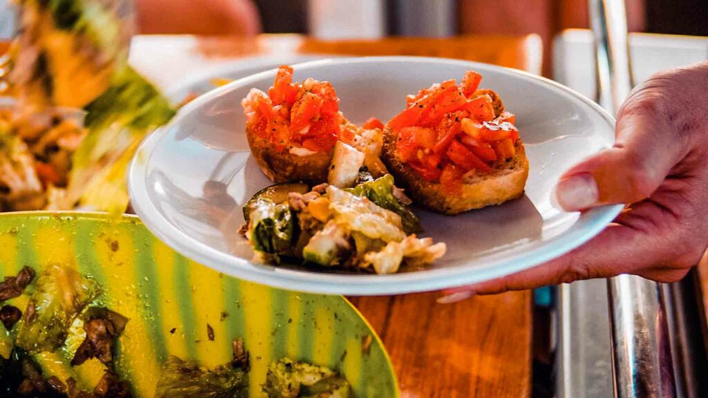 Eine Hand präsentiert einen Teller mit Vorspeisen, darunter zwei Bruschettas mit Tomatenwürfeln und ein weiteres Gericht mit geröstetem Gemüse, in einem lebhaften, zwanglosen Speiseambiente auf einer Segelyacht.
