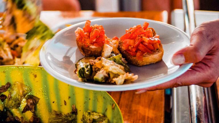 Eine Hand präsentiert einen Teller mit Vorspeisen, darunter zwei Bruschettas mit Tomatenwürfeln und ein weiteres Gericht mit geröstetem Gemüse, in einem lebhaften, zwanglosen Speiseambiente auf einer Segelyacht.