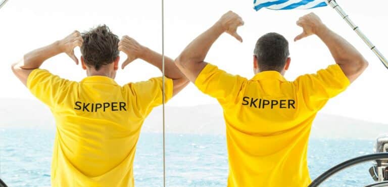 Zwei Männer in gelben „Skipper“-Hemden stehen am Steuer einer Segelyacht und blicken auf das Meer, wobei einer von ihnen das Steuerrad des Schiffs festhält.