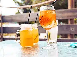 Zwei erfrischende Getränke auf einem Tisch im Freien während eines Segelurlaubs: ein großes Glas Eistee mit Strohhalm und ein Becher Orangensaft vermitteln eine entspannte, sommerliche Stimmung.