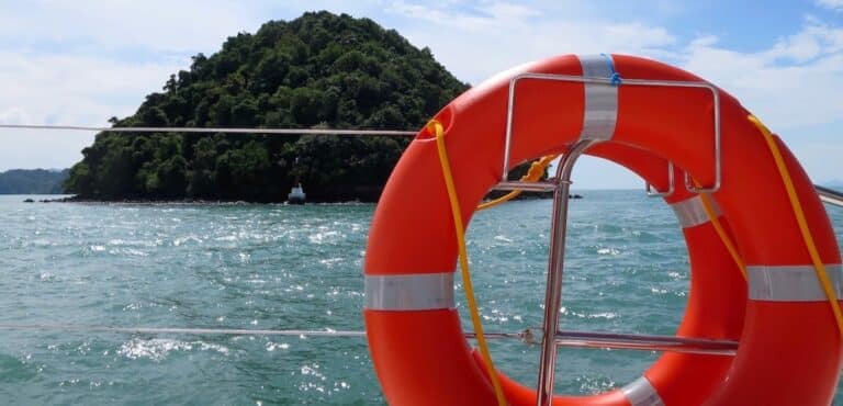 Ein orangefarbener Rettungsring, der an der Reling einer Segelyacht befestigt ist, mit einer üppig grünen Insel und dem blauen Meer im Hintergrund unter einem teilweise bewölkten Himmel.