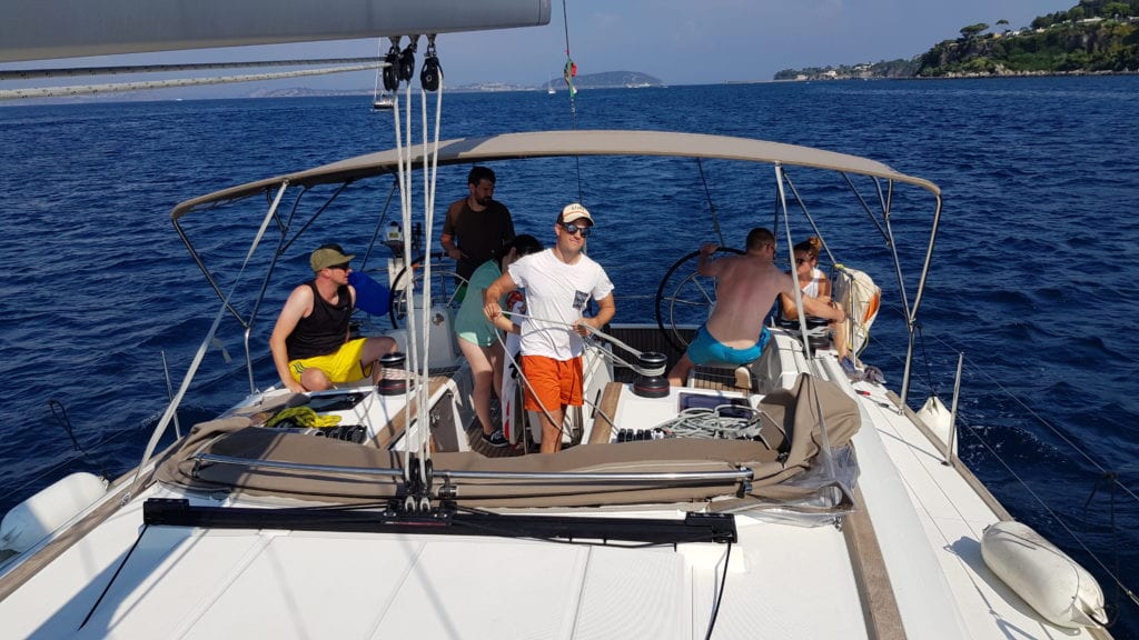 Eine Gruppe von Menschen genießt einen sonnigen Tag auf einem Segelboot mit klarem blauen Himmel und ruhiger See um sie herum. Einige sitzen und einer steht und steuert das Boot während des Segeltörns.