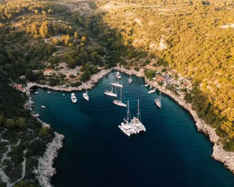 Luftaufnahme einer ruhigen Bucht mit Segelyachten, die im klaren blauen Wasser vertäut sind, umgeben von einer zerklüfteten Küste und dichten grünen Wäldern.