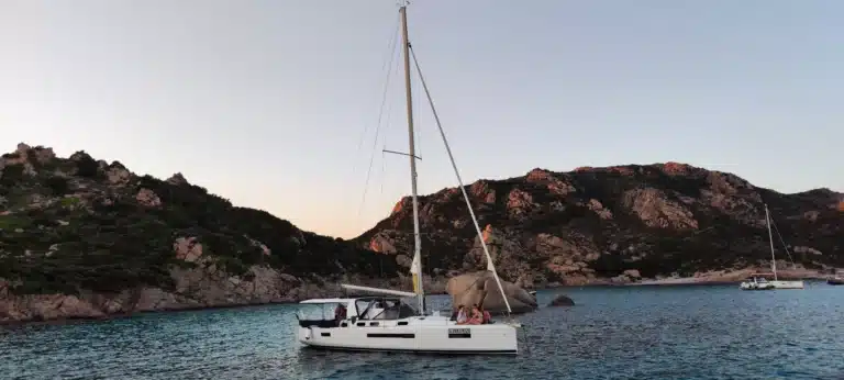 Ein ruhiger Blick auf eine weiße Segelyacht, die in der Abenddämmerung auf dem ruhigen Meer in der Nähe felsiger, baumbestandener Küsten schwimmt. Im Hintergrund sind unter einem zartrosa Himmel andere Boote zu sehen.