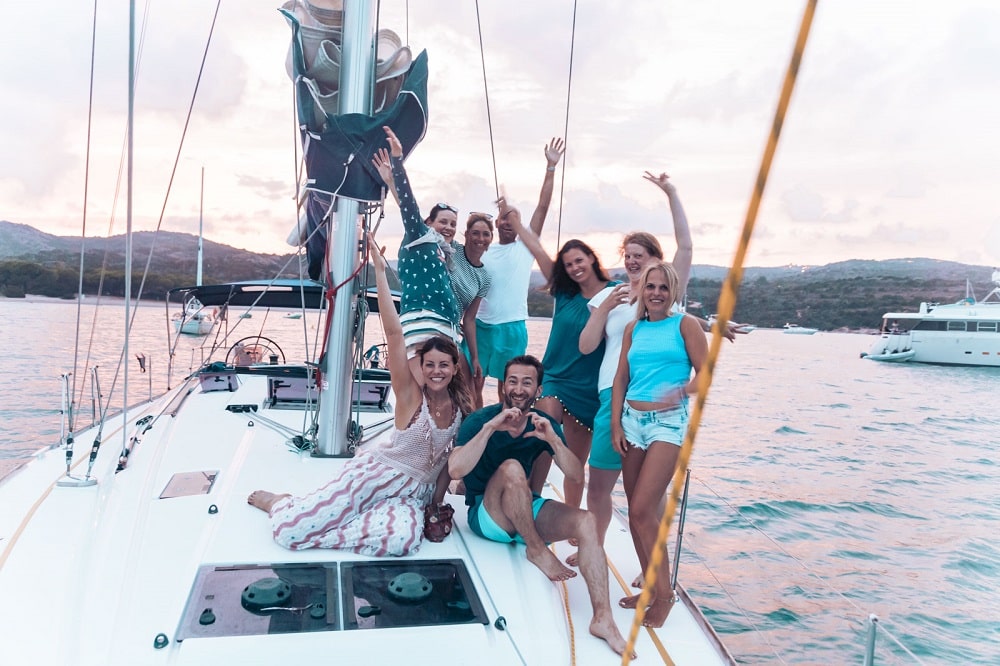 Eine Gruppe von sieben Freunden genießt einen freudigen Moment auf einer Segelyacht, winkt und posiert für ein Foto mit einem malerischen Sonnenuntergang über dem Wasser im Hintergrund. Drei sitzen, während vier stehen,