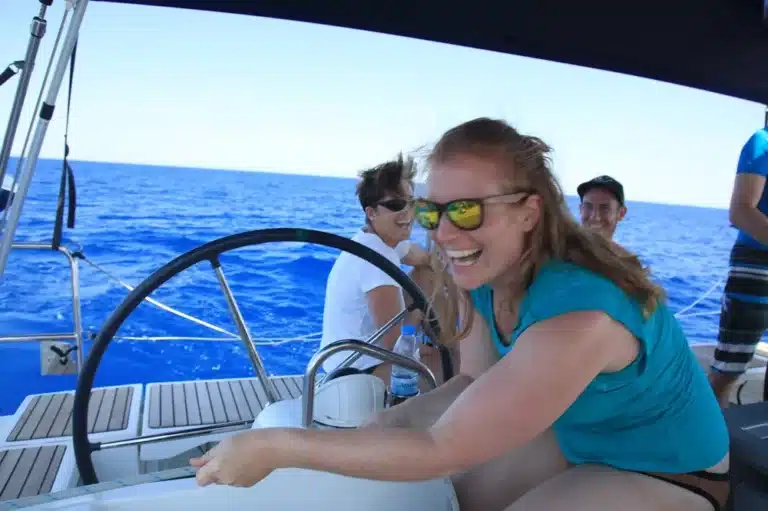 Eine Frau mit roten Haaren, Sonnenbrille und blaugrünem Oberteil steuert fröhlich eine Segelyacht. Im Hintergrund sind weitere Menschen und das offene Meer zu sehen.