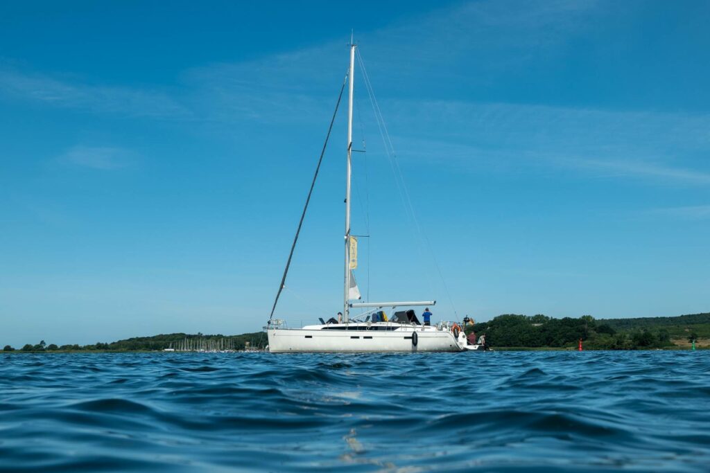Eine Segelyacht mit einem ungesetzten Segel auf ruhigem blauen Wasser, unter einem klaren Himmel, mit sichtbaren Personen auf dem Achterdeck.