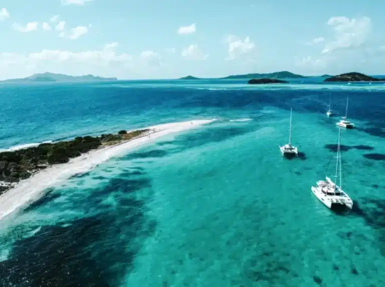 Luftaufnahme eines schmalen Sandstrands mit kristallklarem, türkisfarbenem Wasser, umgeben von mehreren Segelyachten vor einer üppig grünen Insel.