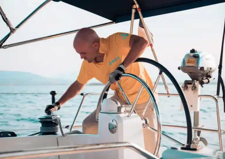 Ein Mann im gelben Hemd steuert in seinem Segelurlaub ein Segelboot und lächelt, während er Einstellungen an einem Bedienfeld vornimmt, im Hintergrund das Meer.