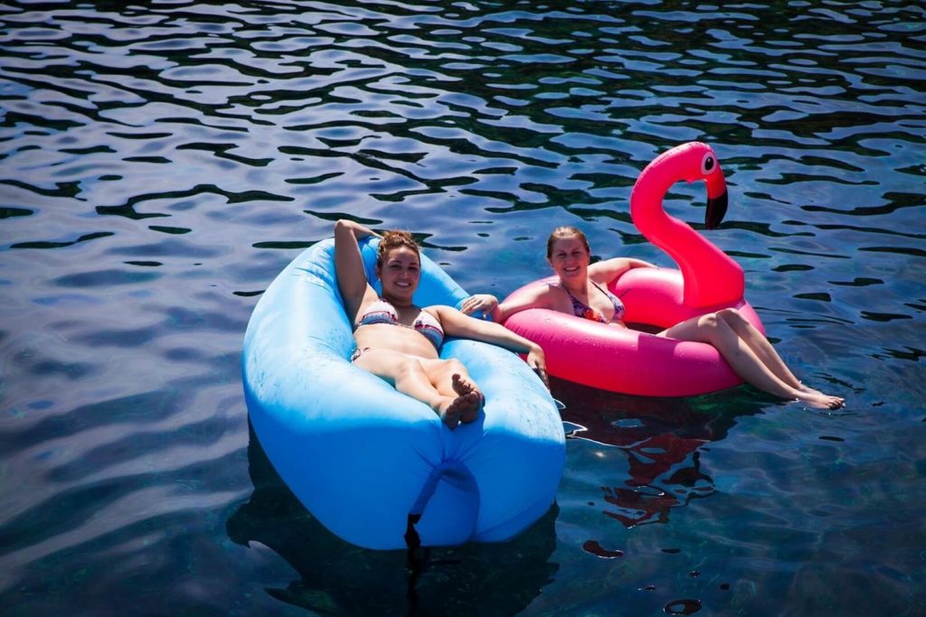 Zwei Menschen entspannen auf aufblasbaren Schwimmkörpern, einer in Form eines Flamingos und der andere in Form eines blauen Rings, und segeln an einem hellen Tag im klaren, blauen Wasser.