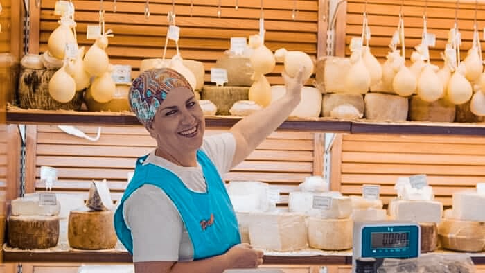 Eine lächelnde Frau mit Kopftuch und Schürze zeigt auf hängende Käsesorten an einem Marktstand, hinter ihr sind während ihres Segelurlaubs zahlreiche Käselaibe auf Regalen ausgestellt.