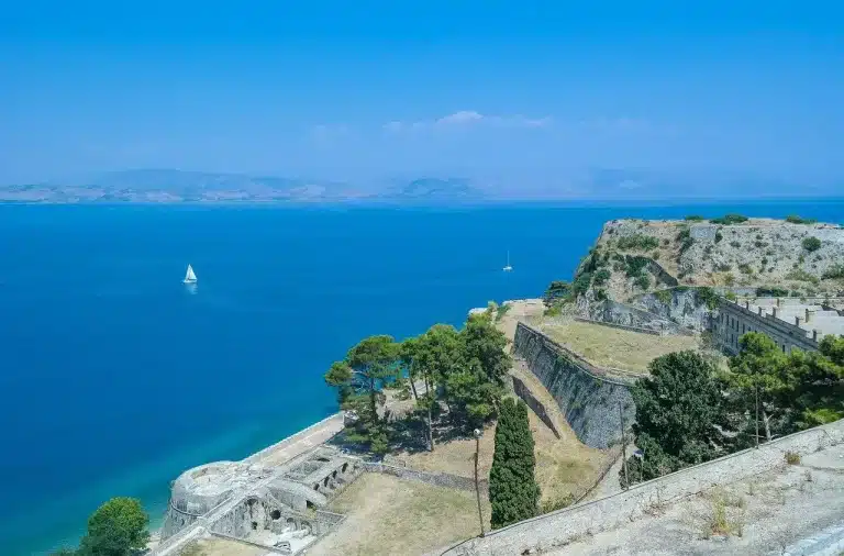 Blick auf eine alte Steinfestung mit Blick auf ein klares blaues Meer mit Segelbooten und fernen Hügeln unter einem strahlend blauen Himmel, perfekt für einen Segelurlaub.