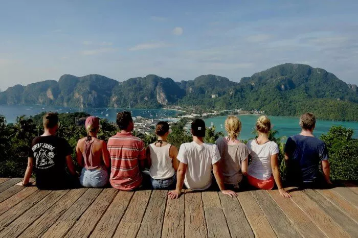 Sieben Personen sitzen auf einer Holzplattform und genießen während eines Segeltörns eine malerische Aussicht auf eine üppige Küstenregion mit Bergen und klarem blauen Himmel.