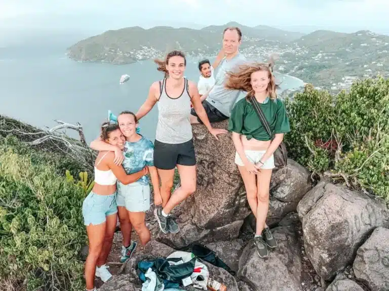 Eine fröhliche fünfköpfige Familie posiert auf einem felsigen Hügel mit einem malerischen Blick auf die Küste im Hintergrund. Der Wind zerzaust ihre Haare, während sie für das Foto lächeln und einen perfekten Moment während ihres Segens festhalten.