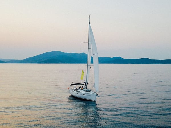 Ein kleines Segelboot mit weißem Segel gleitet bei Sonnenuntergang über ruhige Gewässer. Im Hintergrund sind sanfte Hügel unter einem zartrosa Himmel zu sehen. Genießen Sie ein unvergessliches „Segelyacht“-Erlebnis!