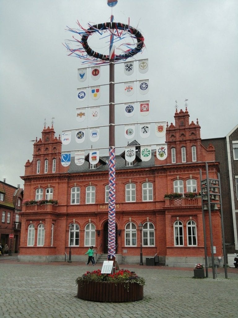 Ein traditioneller Maibaum mit bunten Bändern und verschiedenen Wappen, der während eines Segeltörns unter einem bewölkten Himmel vor einem roten Backsteingebäude auf einem europäischen Stadtplatz aufgestellt wird.