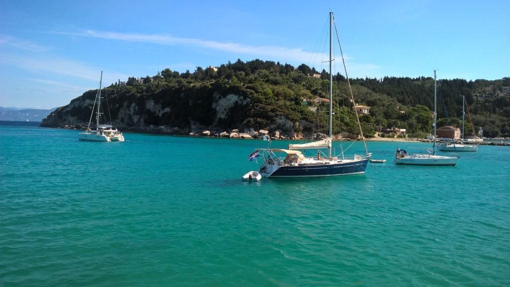 Segelyachten, die im klaren, türkisfarbenen Wasser in der Nähe eines grün bewachsenen Hügels unter einem klaren, blauen Himmel vor Anker liegen.