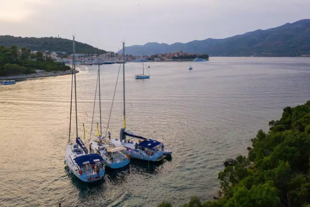 Luftaufnahme von drei Segelyachten, die in Küstennähe vor Anker liegen, mit einer ruhigen Bucht und einer kleinen Stadt zwischen Hügeln im Hintergrund in der Abenddämmerung.