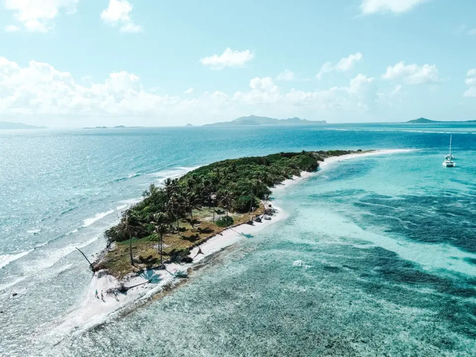 Luftaufnahme einer tropischen Halbinsel mit üppigem Grün, umgeben von klarem, türkisfarbenem Wasser, mit einem Sandstrand und einer Segelyacht in der Nähe.