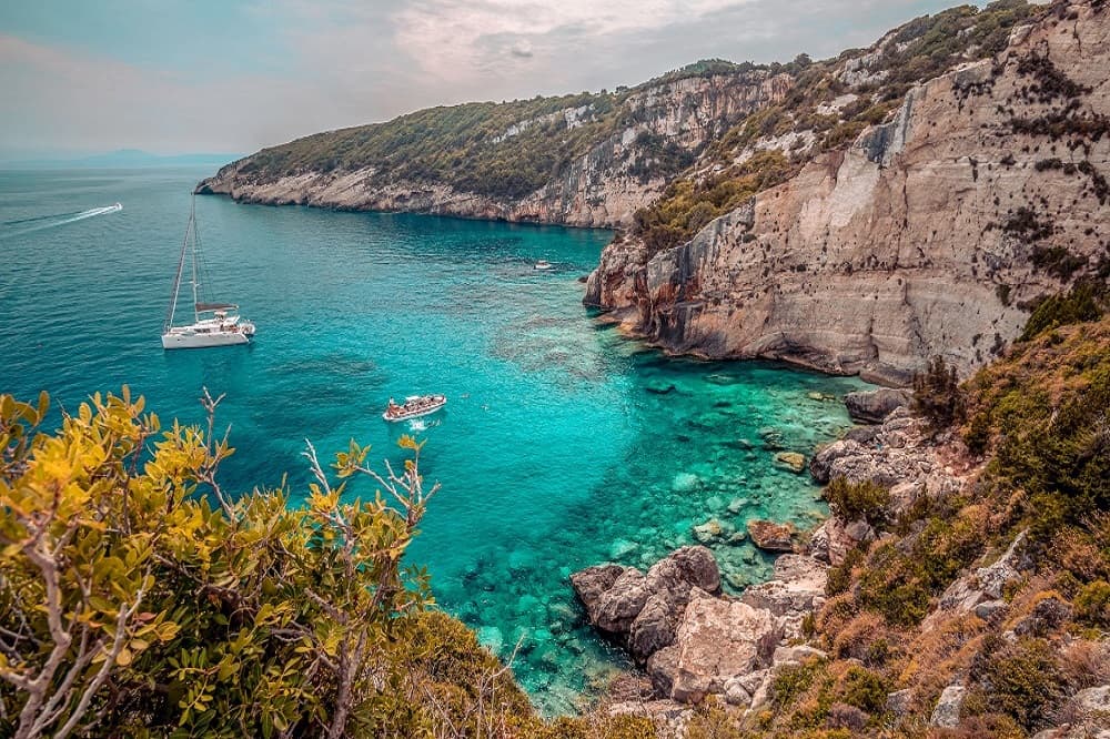 Eine atemberaubende Küstenszene mit türkisfarbenem Wasser, umgeben von steilen, felsigen Klippen. Zwei weiße Segelboote schwimmen in Ufernähe und üppiges Grün ziert die Ränder.
