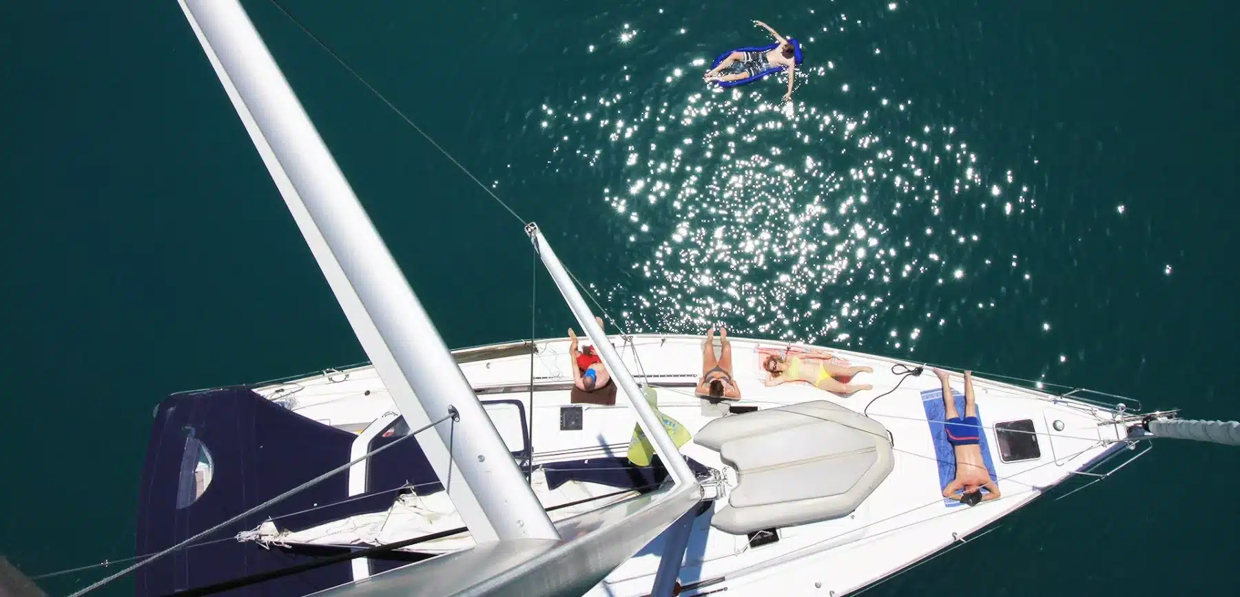 Luftaufnahme einer Segelyacht auf klarem Wasser mit vier Personen, die auf dem Deck ein Sonnenbad nehmen, und einer Person, die in der Nähe auf einem Schlauchboot schwimmt.