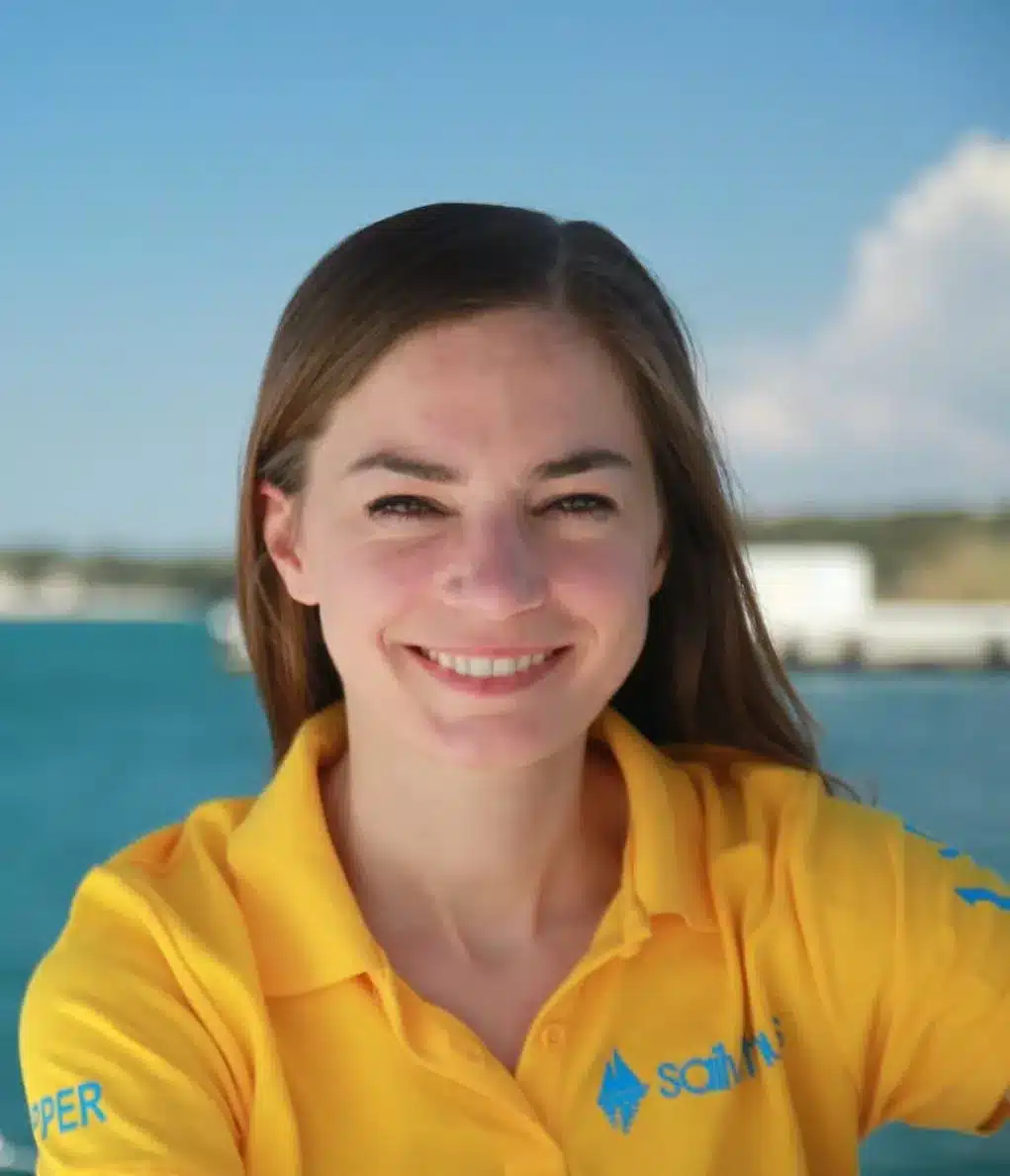 Eine lächelnde Frau in einem gelben Poloshirt mit einem Logo darauf steht vor einem hellblauen Segelyacht-Hintergrund.