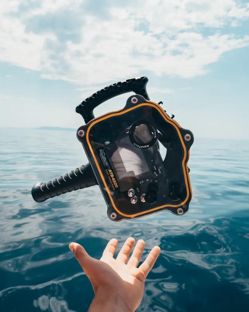 Während eines ruhigen Segeltörns unter einem klaren blauen Himmel streckt sich eine Hand aus, um ein schwimmendes Unterwasserkameragehäuse vor der ruhigen Meereskulisse einzufangen.