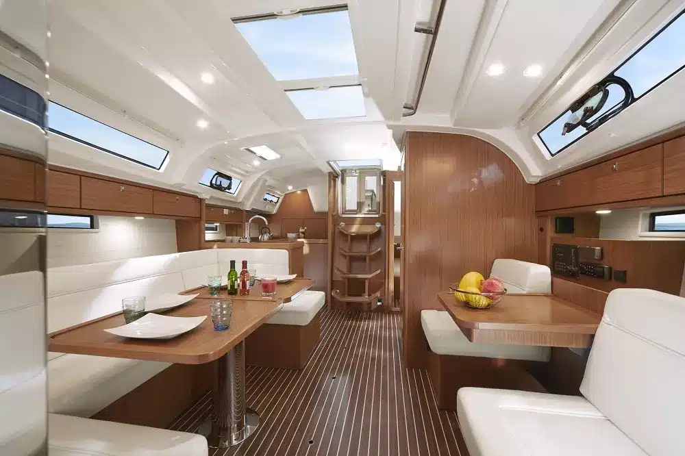 Innenraum einer Luxusyachtkabine mit Holzverkleidung, mit einem Essbereich mit weißen gepolsterten Bänken und einem zentralen Tisch, ideal für Segelurlaub, ergänzt durch eine Küchenzeile und eine