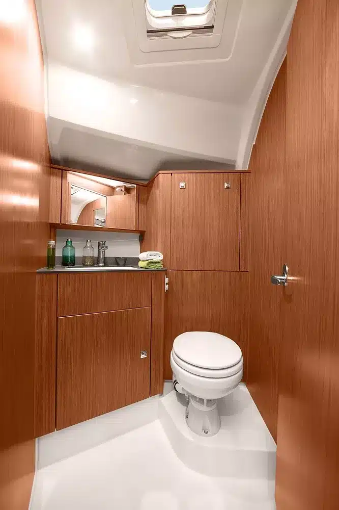 Ein modernes kleines Badezimmer in einem Katamaran mit glänzend weißen Böden, Holzschränken und einer kompakten Toilette, in das durch eine Luke oben Tageslicht einfällt.