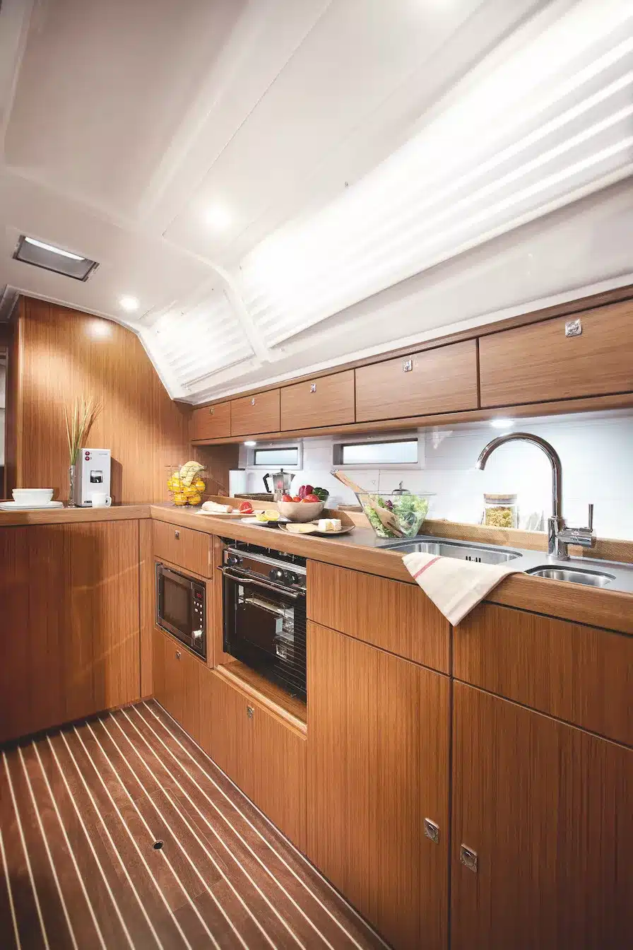 Innenansicht einer modernen Segelyacht-Küche mit eleganten Holzschränken, Geräten aus Edelstahl und einer ordentlich organisierten Arbeitsfläche. Natürliches Licht durchflutet den Raum durch die Luken an der Decke.