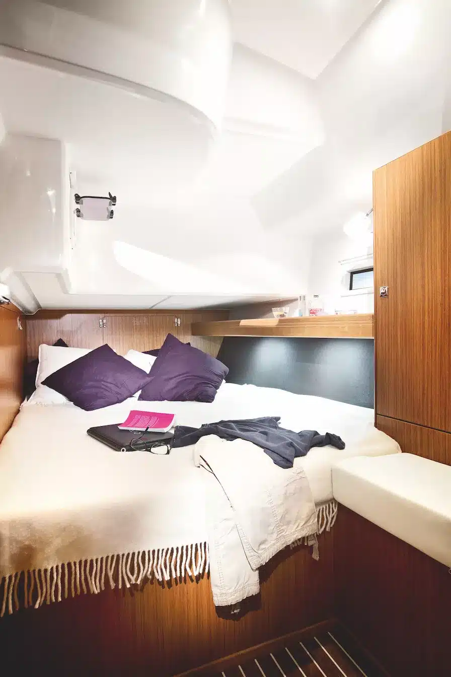 Innenansicht einer aufgeräumten Segelyachtkabine mit einem ordentlich gemachten, von Holzvertäfelung umgebenen Bett, violetten Kissen, einem Buch und lässig auf dem Bett platzierter Kleidung.