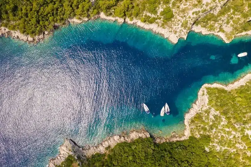 Luftaufnahme einer kristallklaren blauen Bucht mit drei Segelyachten, die in der Nähe der felsigen Küste vor Anker liegen und von üppigem Grün umgeben sind.