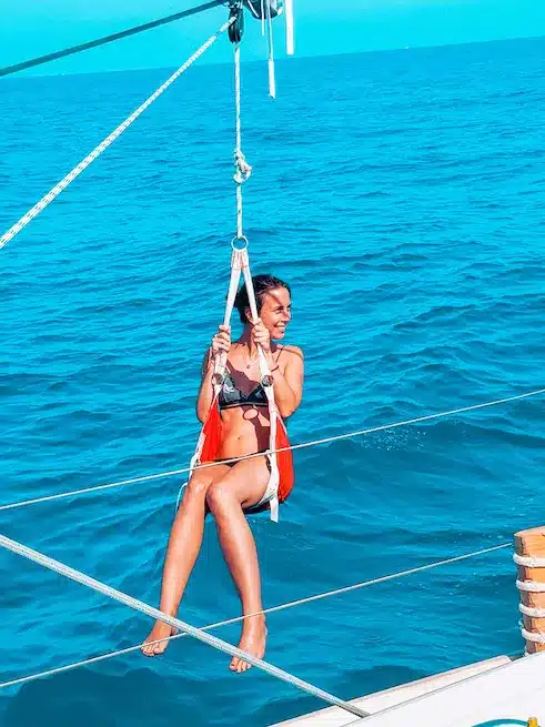Eine Frau in einem roten Badeanzug schaukelt auf einer an einer Segelyacht befestigten Seilschaukel und genießt an einem sonnigen Tag den blauen Ozean im Hintergrund.