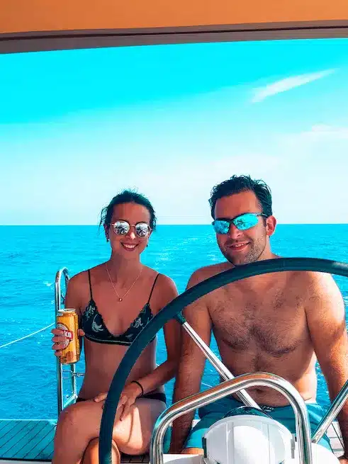 Ein Mann und eine Frau genießen einen sonnigen Tag auf einem Boot während ihres Segeltörns, wobei der Mann steuert. Die Frau hält ein Getränk in der Hand, beide lächeln und tragen Sonnenbrillen, mit klarem blauen