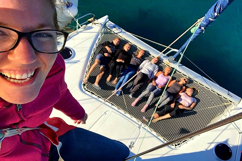 Eine lächelnde Frau macht ein Selfie auf einer Segelyacht, im Hintergrund sieht man eine Gruppe von vier Freunden, die auf einem Netz über dem klaren blauen Wasser faulenzen.