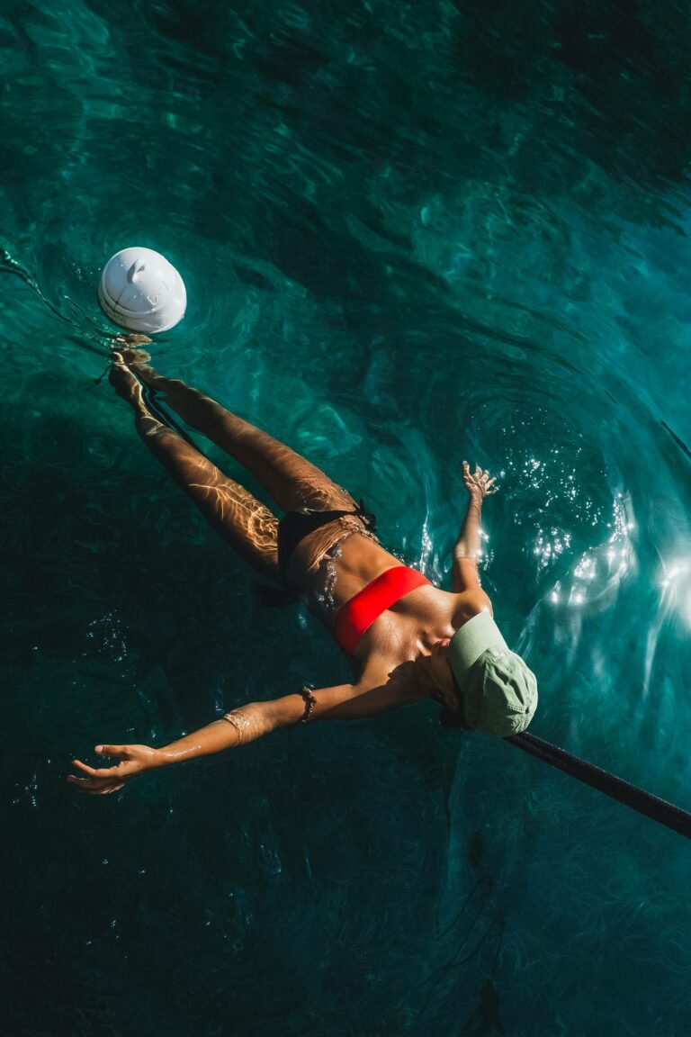 Eine Person in einem roten Bikinioberteil und einer grünen Hose schwimmt während eines Segeltörns im klaren blauen Wasser und versucht, einen weißen Ball zu fangen. Sonnenlicht erzeugt schimmernde Muster auf der Oberfläche.