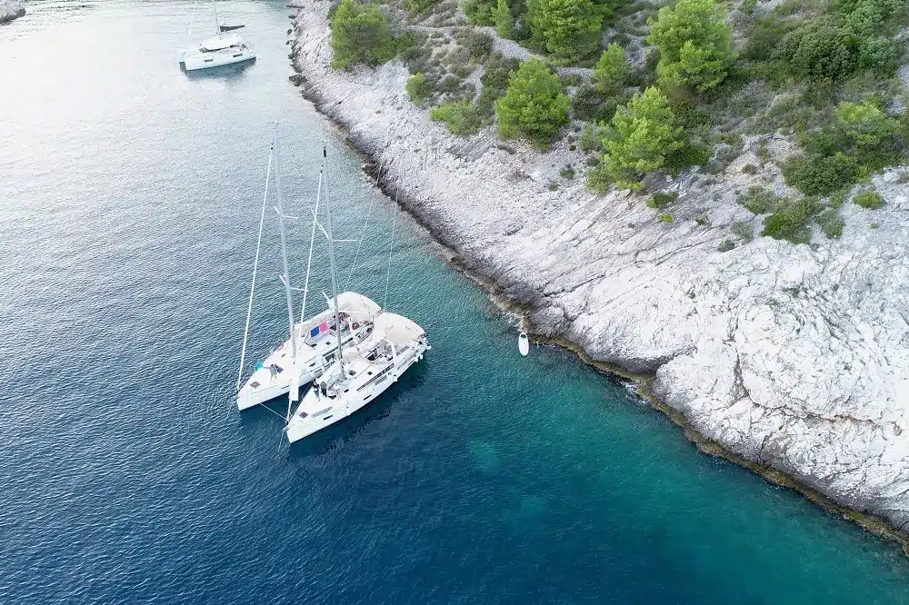 Luftaufnahme von zwei Segelbooten, die nahe der felsigen Küste eines klaren, blauen Meeres vor Anker liegen, umgeben von üppigem Grün, perfekt für einen Segelurlaub.