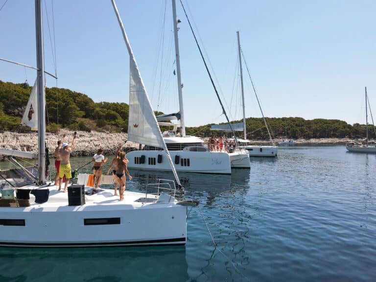 Menschen genießen das sonnige Wetter, indem sie auf einer angedockten weißen Segelyacht winken und Fotos machen, umgeben von anderen Yachten auf ruhiger See in der Nähe einer felsigen, mit Grün bedeckten Küste.