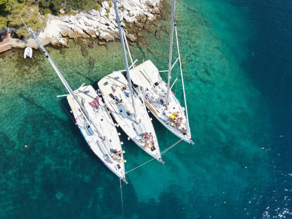 Luftaufnahme von drei Segelbooten, die dicht beieinander in der Nähe einer felsigen Küste im klaren, türkisfarbenen Wasser vertäut sind. Auf den Decks sind Menschen zu sehen, die ihren Segelurlaub genießen.