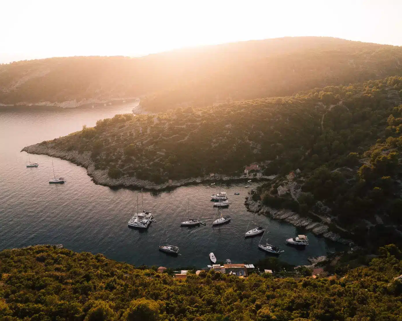 Luftaufnahme einer ruhigen Bucht bei Sonnenuntergang mit Segelyachten, die in der Nähe einer waldbedeckten Küste vor Anker liegen und vom goldenen Sonnenlicht erhellt werden.