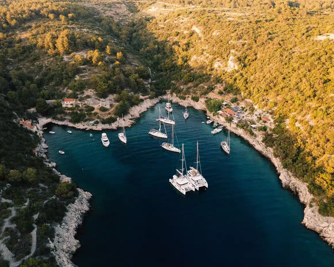 Luftaufnahme einer ruhigen Bucht mit türkisfarbenem Wasser, umgeben von einem üppigen grünen Wald, mit Yachten und Booten, die während ihres Segelurlaubs in der Nähe der zerklüfteten Küste vor Anker liegen.