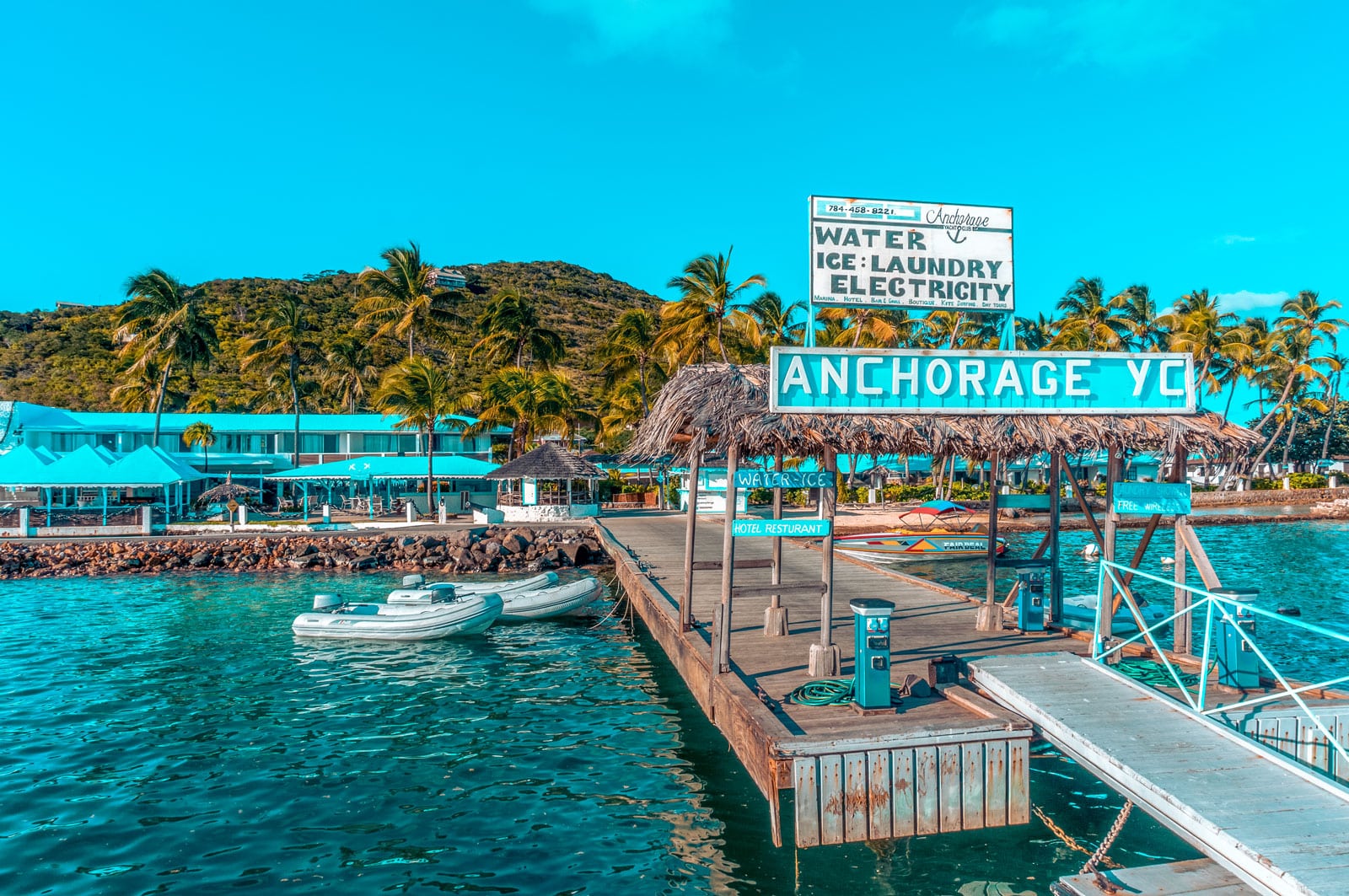 Ein helles, farbenfrohes Bild des Piers des Anchorage Yacht Clubs mit einem Schild mit den Annehmlichkeiten, daneben liegt eine kleine Segelyacht vor Anker, unter einem klaren blauen Himmel. Tropische Szene mit üppigem Grün im Hintergrund.