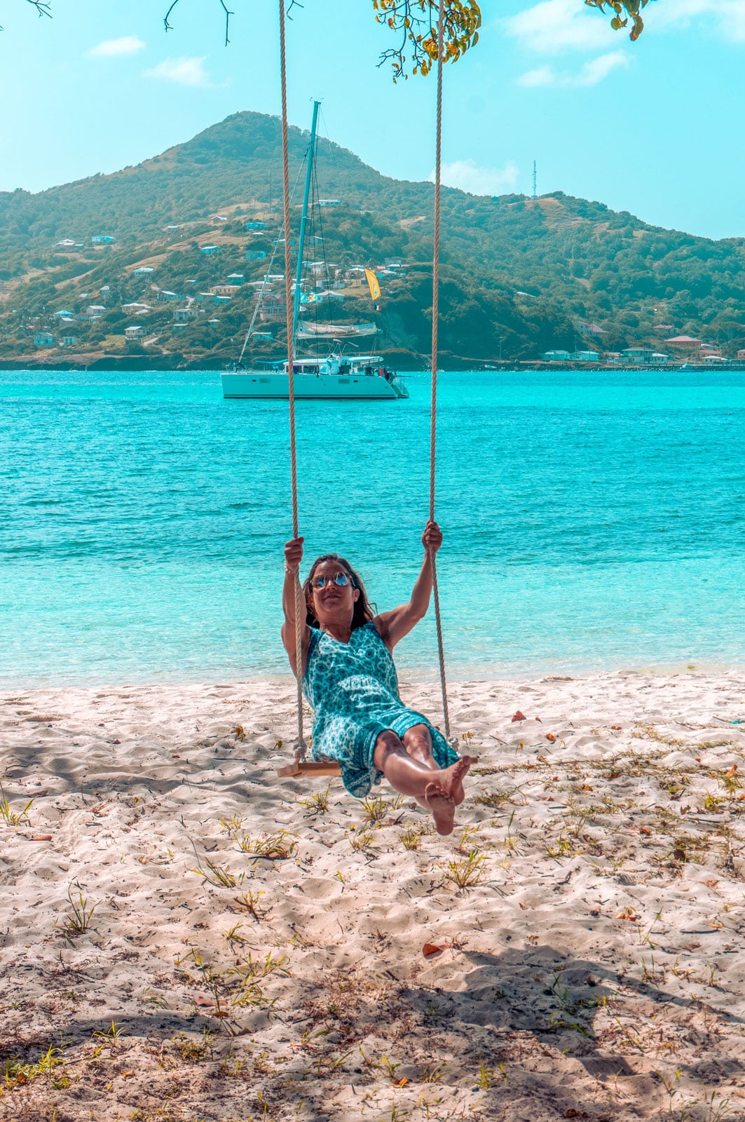 Eine Frau schaukelt während ihres Segelurlaubs auf einer Seilschaukel an einem tropischen Strand und lächelt in die Kamera vor der malerischen Kulisse des klaren blauen Wassers und einer bergigen Insel.