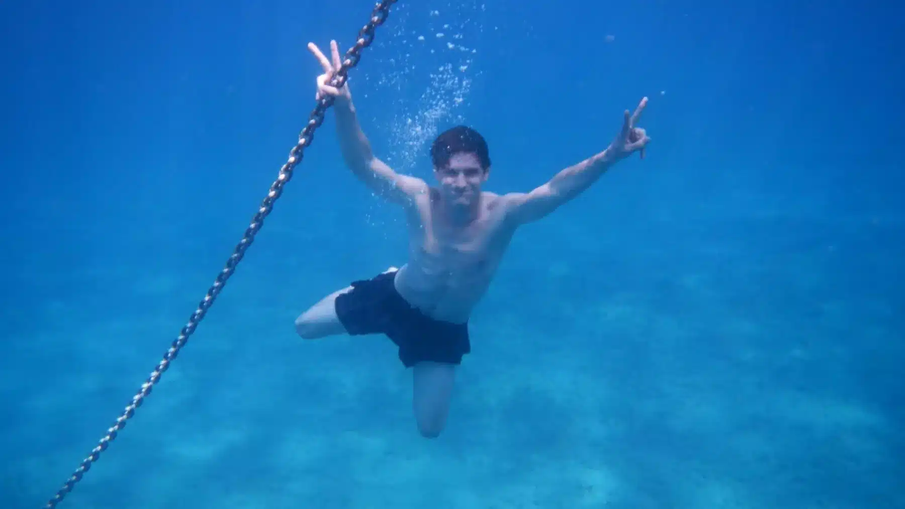 Ein Mann unter Wasser, der freudig beide Arme in einem V-Zeichen ausstreckt, schwimmt neben einer nach unten gerichteten Kette, aufgenommen vor einem klaren blauen Wasserhintergrund in der Nähe einer Segelyacht.