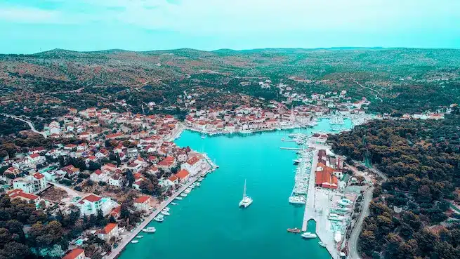 Luftaufnahme einer Küstenstadt mit Gebäuden mit roten Dächern, üppigem Grün und Segelyachten, die in einem türkisfarbenen Hafen vor Anker liegen.