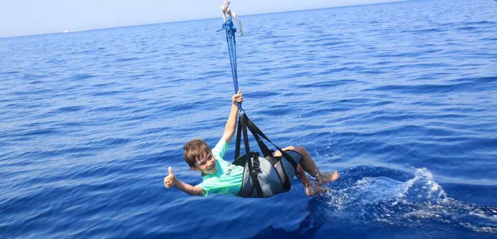 Eine Person in einem grünen Hemd und Shorts fliegt mit dem Parasailing über den klaren blauen Ozean und streckt den Daumen nach oben, während sie in einem an einer Segelyacht befestigten Gurt hängt.