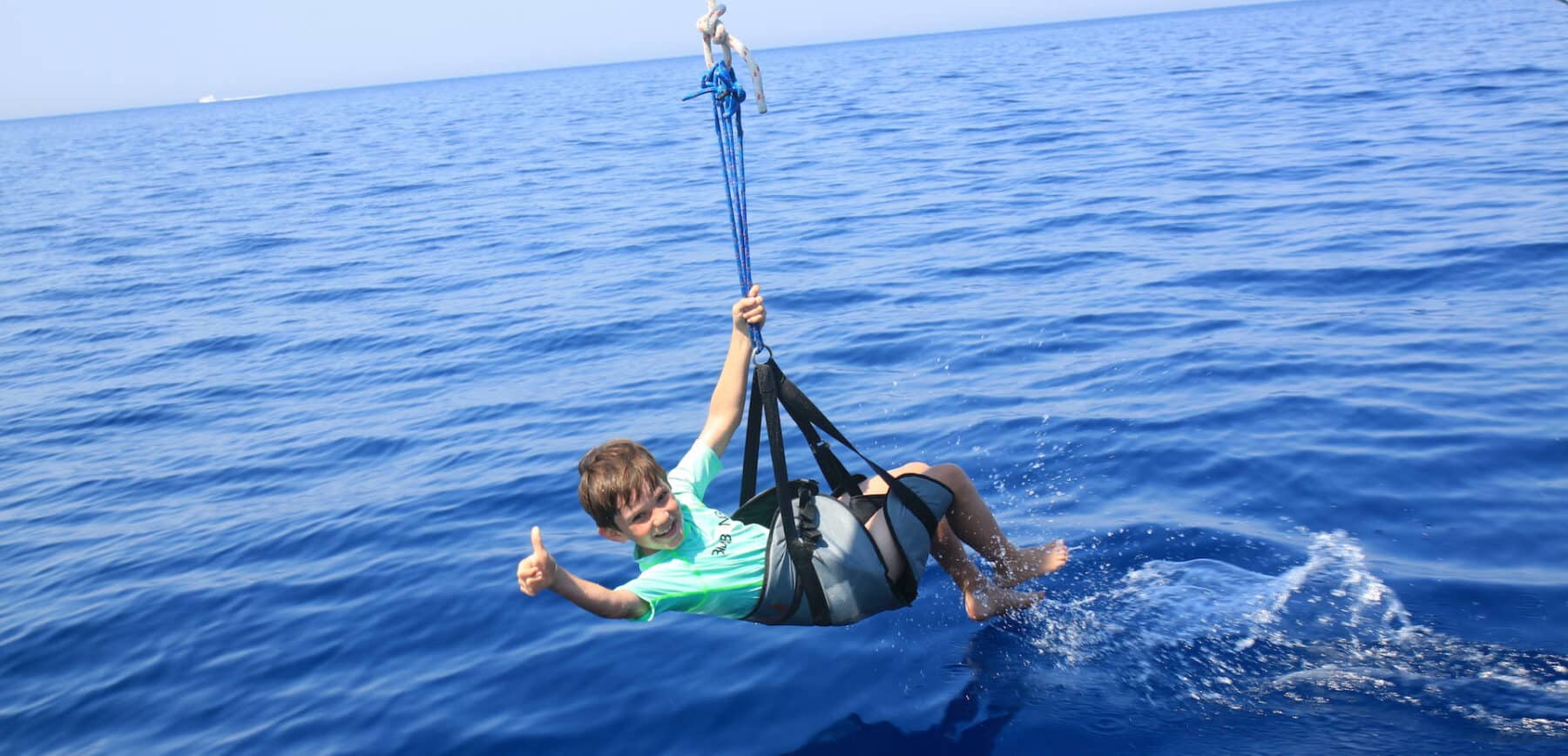 Eine Person in einem grünen Hemd und Shorts fliegt mit dem Parasailing über den klaren blauen Ozean und streckt den Daumen nach oben, während sie in einem an einer Segelyacht befestigten Gurt hängt.