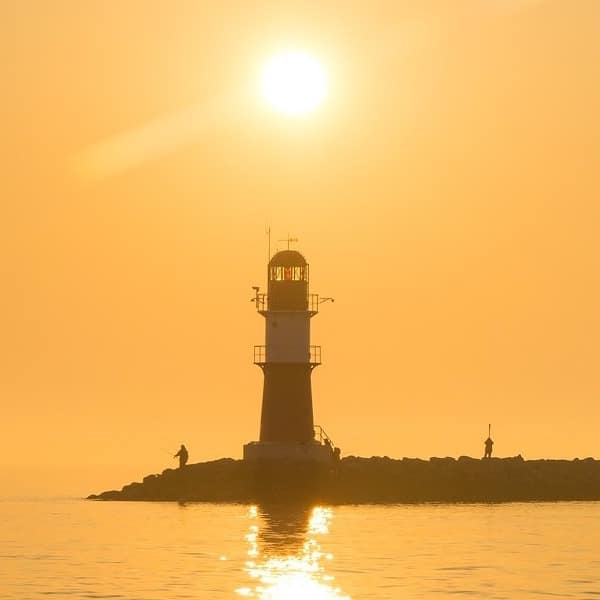 Die Silhouette eines Leuchtturms und zweier Menschen auf einem Felsvorsprung, im Gegenlicht eines strahlend goldenen Sonnenaufgangs, der sich während einer Segelreise im Wasser spiegelt.